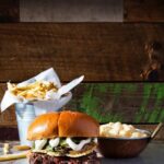 A burger sits on a table in front of a container of fries and bowl of mashed potatoes.