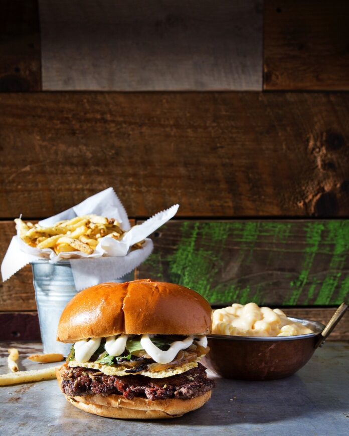 A burger sits on a table in front of a container of fries and bowl of mashed potatoes.