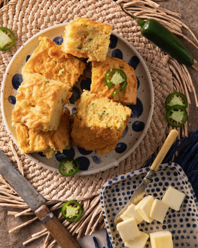 A white plate with blue patterns sits on a table with pieces of cheddar cornbread piled high and a few jalepenos scattered throughout. Nearby sits a plate of butter slices and a knife for spreading.