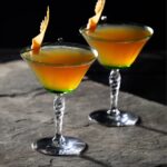 Two cocktail glasses hold the Dance of the Dragons House of Dragons cocktail in an orange-ish color with a manicured grapefruit sticking out of each glass on a stone table.