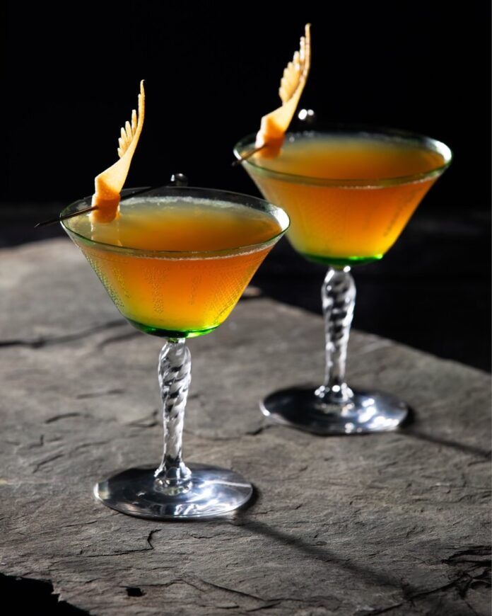 Two cocktail glasses hold the Dance of the Dragons House of Dragons cocktail in an orange-ish color with a manicured grapefruit sticking out of each glass on a stone table.