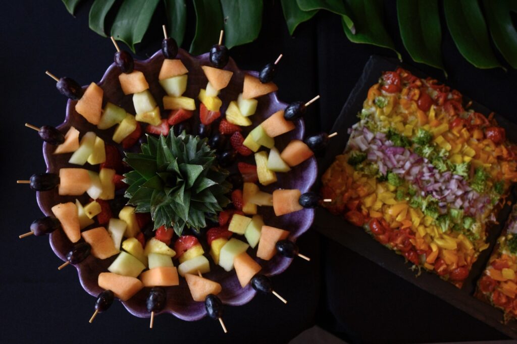 A bowl of fruit skewers in a circle sits next to a rainbow pizza topped with chopped fresh vegetables.