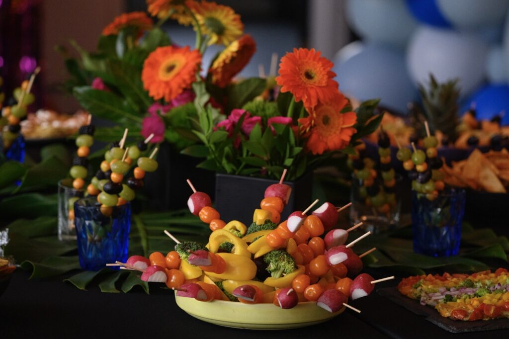 A green bowl full of vegetable skewers sitting on a table amongst florals and fruit skewers in various cups.