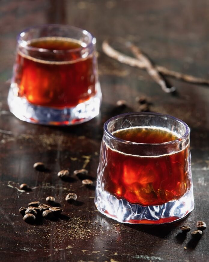 Two short cocktail glasses hold a red Fury is Born cocktail inspired by Mad Max, with coffee beans sitting between the two glasses.