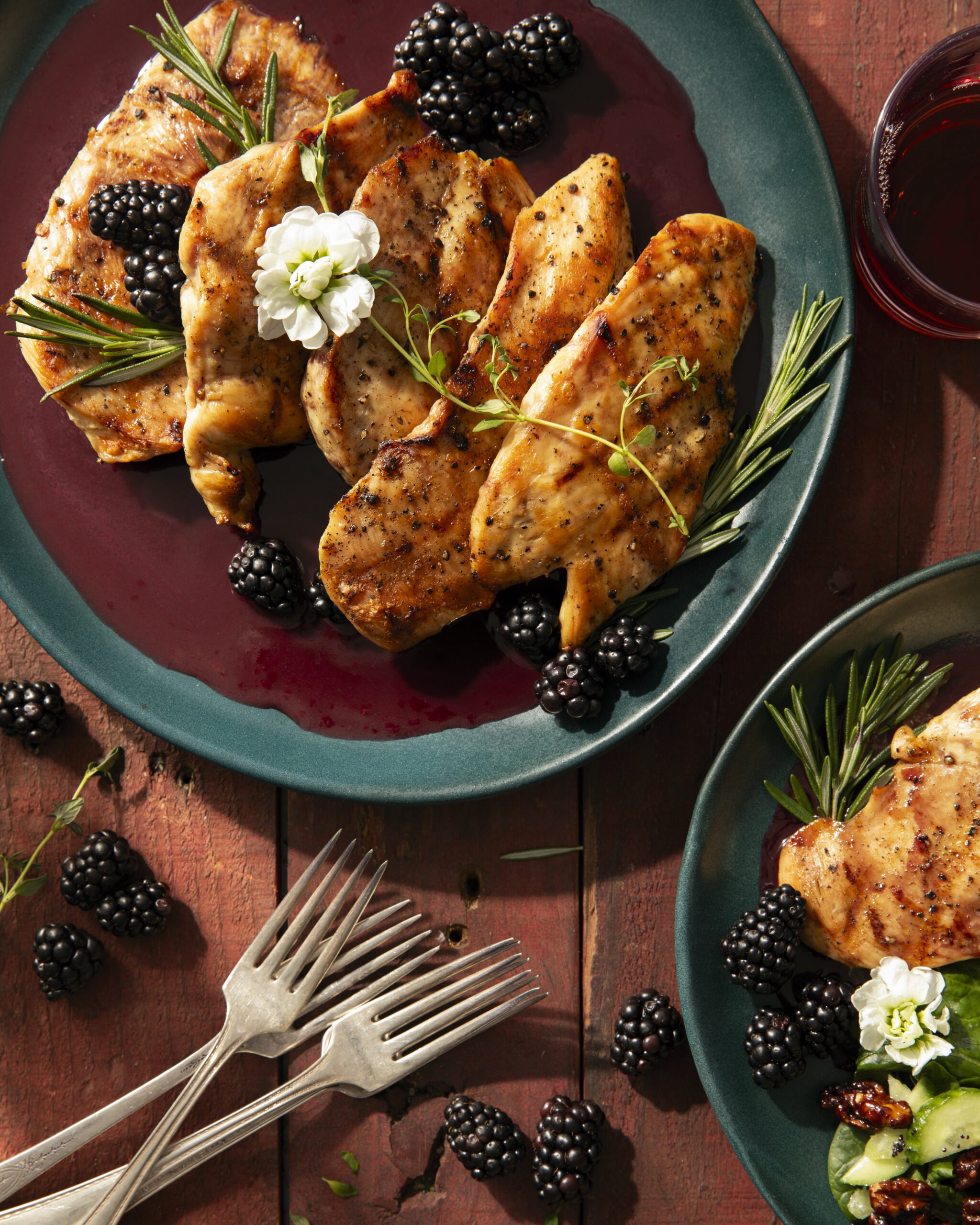 Grilled chicken breasts garnished with fresh blackberries, rosemary sprigs, and edible flowers, served on a teal plate with a rich, dark blackberry-herb sauce.