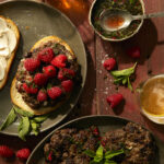 Grilled lamb burgers served on slices of toasted sourdough bread, topped with fresh raspberries, mint leaves, and a raspberry-mint vinegar drizzle, with a glass of beer and extra sauce on the side.