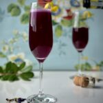 Two champagne flutes of a dark purple Lord & Lady Bridgerton cocktail with a lemon peel twist garnish, butterfly pea flowers below, and a bottle of champagne pouring into the background glass. It's all set against a blue floral background.