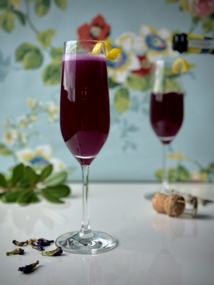 Lord & Lady Bridgerton Cocktail Two champagne flutes of a dark purple Lord & Lady Bridgerton cocktail with a lemon peel twist garnish, butterfly pea flowers below, and a bottle of champagne pouring into the background glass. It's all set against a blue floral background.