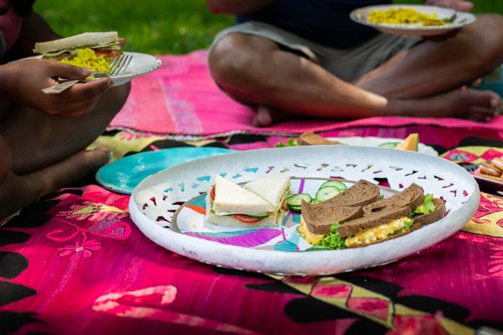 Above a pink picnic blanket sits a white plate with sandwiches on top as a person holds an egg salad sandwich in their hand to the left.