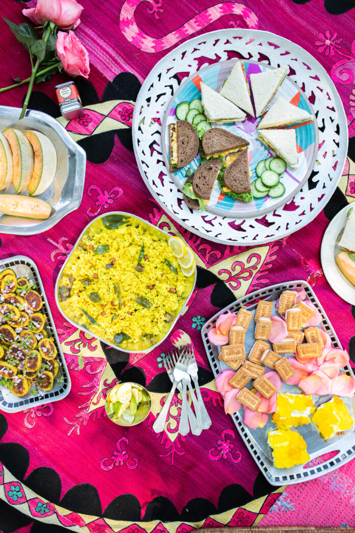 A picnic spread on a pink blanket with plates of yellow lemon rice, rose milk cookies, potato patties, and two different types of sandwiches on a big round plate.