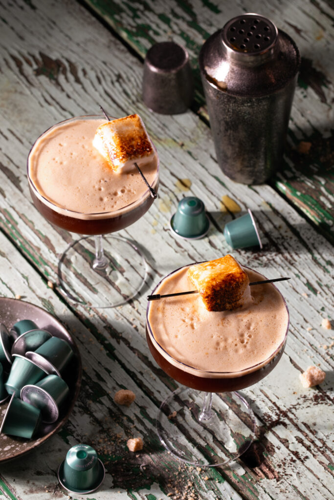 Two Toasted Marshmallow Espresso Martinis in two glasses with Nespresso pods and a cocktail shaker nearby.