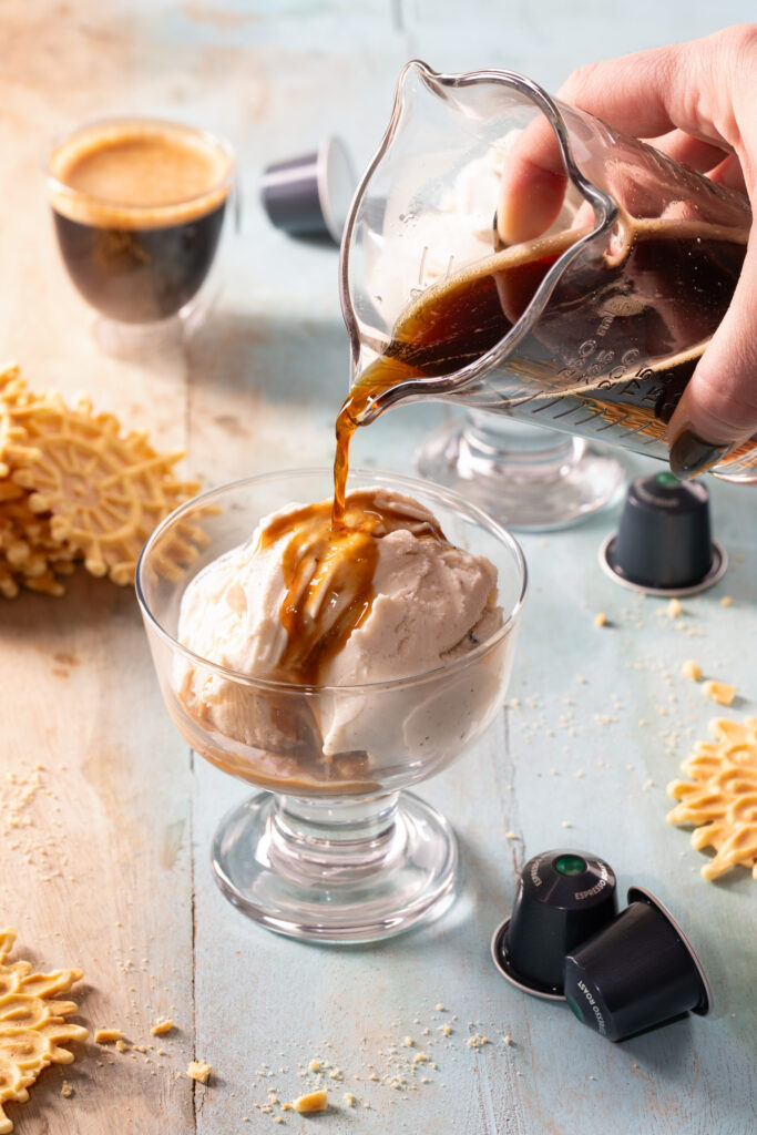 A hand pours a shot of espresso over a cup of ice cream to create a Nespresso Affogato with pizzelles and a shot of espresso in the background.