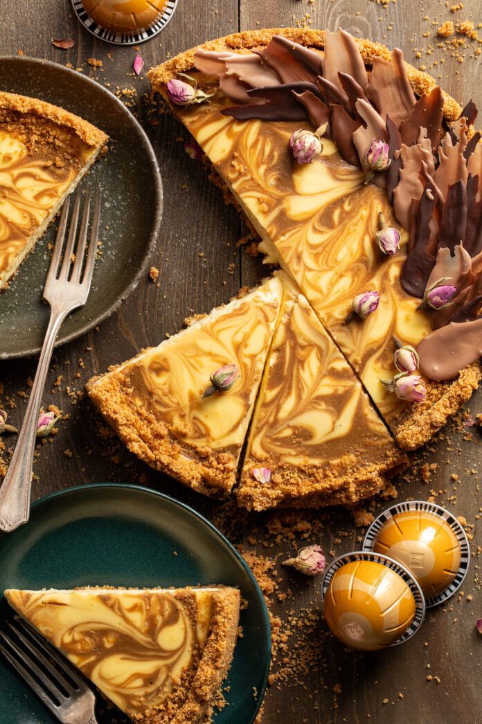 An espresso swirl cheesecake sits on a wooden table with two slices cut out which are displayed on dark green plates with a fork on each.