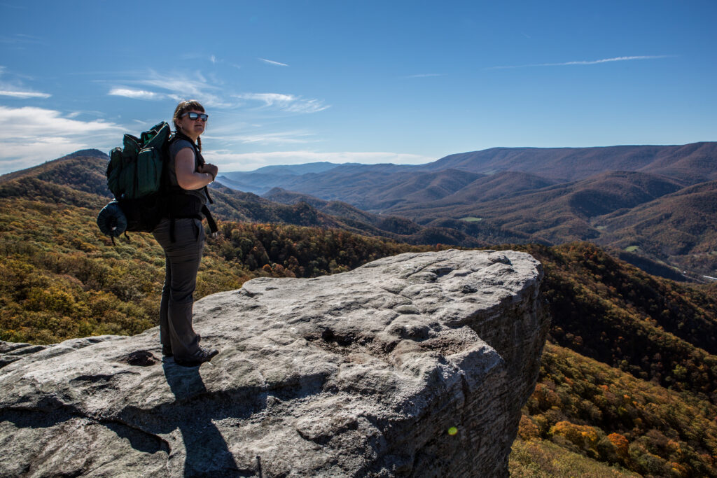 A female backpacker standing at the edge of a cliff with a blue sky and mountains in the background