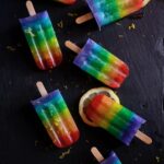 7 rainbow pride popsicles sit on a black table with slices of lemon scattered beneath them.
