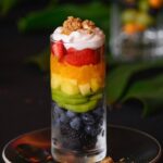 A glass holds various fresh fruits in a rainbow pattern with whipped topping and granola on top, all sitting on top of a black plate beside a spoon.