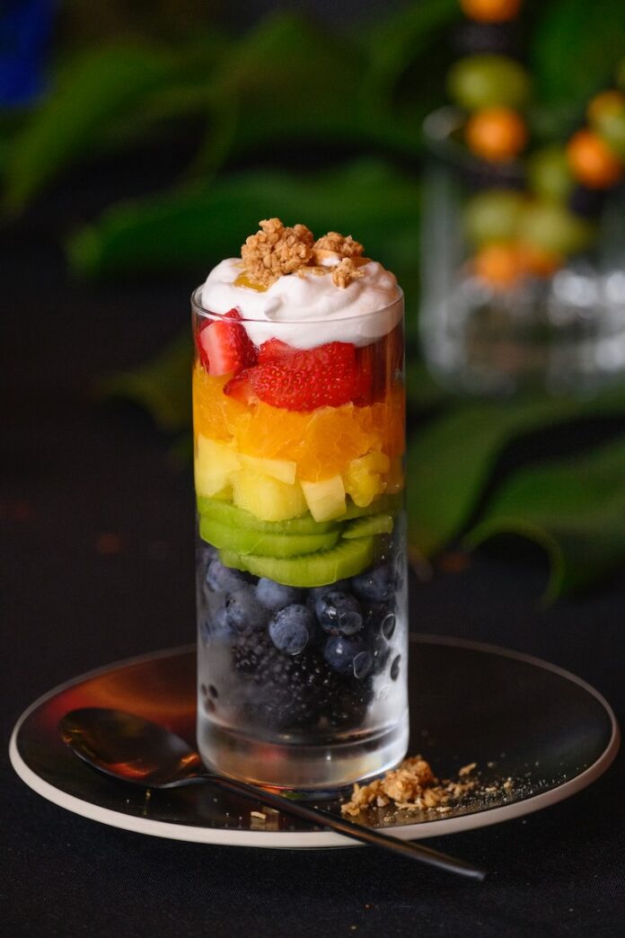 A glass holds various fresh fruits in a rainbow pattern with whipped topping and granola on top, all sitting on top of a black plate beside a spoon.