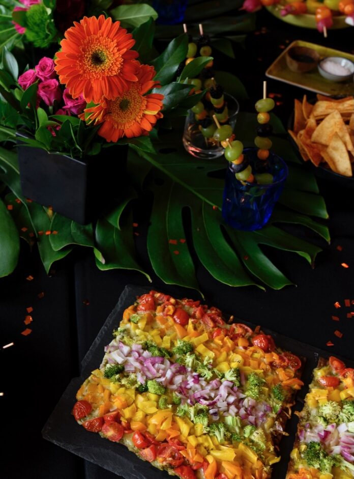 A rainbow flatbread pizza sits below an array of bright colorful flowers with fruit skewers sittin in cups to the right side.