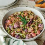 A white bowl full of a Roasted Radish Salad topped with a sprig of dill and featuring more plates, a napkin, and small bowls of seasoning nearby.