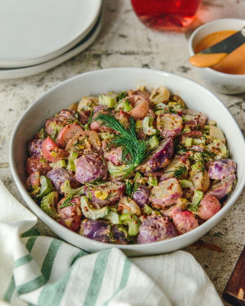 A white bowl full of a Roasted Radish Salad topped with a sprig of dill and featuring more plates, a napkin, and small bowls of seasoning nearby.