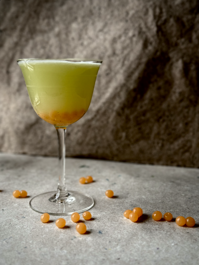 A tall cocktail glass with a green Boba Fett Cocktail and orange boba balls in the bottom of the glass and on the table.