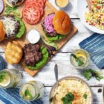 On a wood picnic table sits a wooden board covered in Great Gourmet Burgers with drinks nearby and a bowl of potato salad.