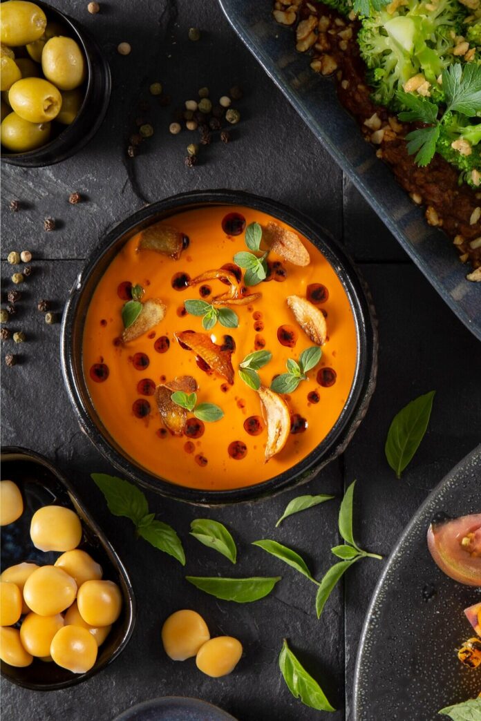 A black bowl holds a red Tomato Gazpacho with vegetable and herb garnishes, surrounded by fresh vegetables and a sheet pan.
