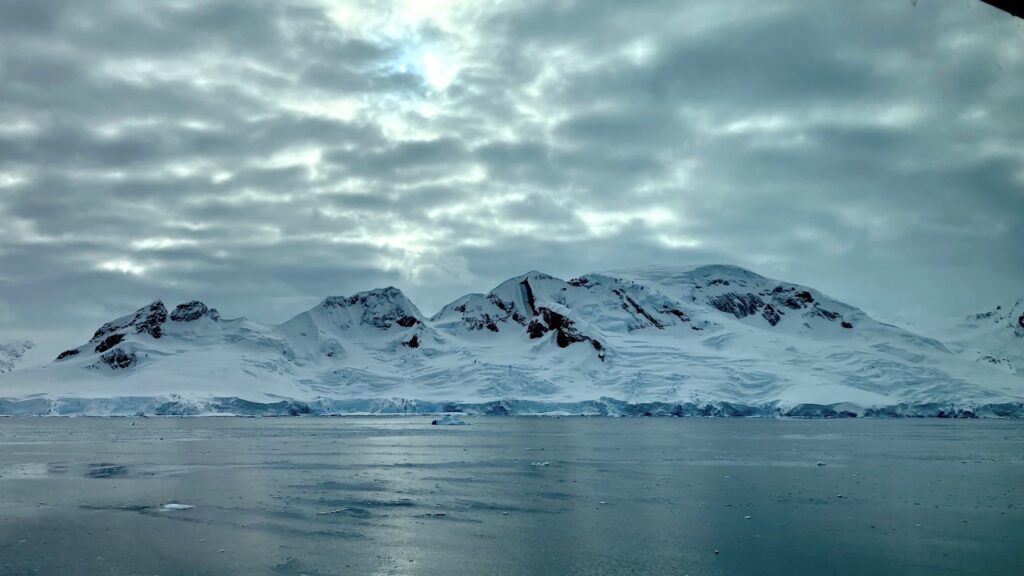 Large peaks of Antarctic mountains sit beneath heavy grey cloudy and above deep grey blue water.