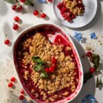 A Sour Cherry Crisp sits in a white dish with a crumble topping as a slice sits on a small white plate nearby.