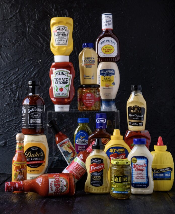 Different brand bottles of condiments in ketchup, mayo, mustard, and beyond sit stacked against a black background.