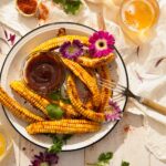 A bowl of corn ribs sits on a wooden table with a small container of homemade BBQ sauce in the center. Two glasses of beer sit in the top right corner and flowers are scattered throughout.