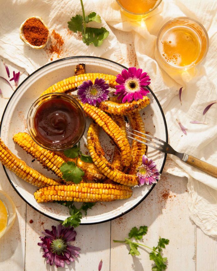 A bowl of corn ribs sits on a wooden table with a small container of homemade BBQ sauce in the center. Two glasses of beer sit in the top right corner and flowers are scattered throughout.