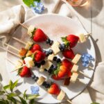 A white plate holds 6 kabobs with strawberries, blueberries, and squares of poundcake on them.