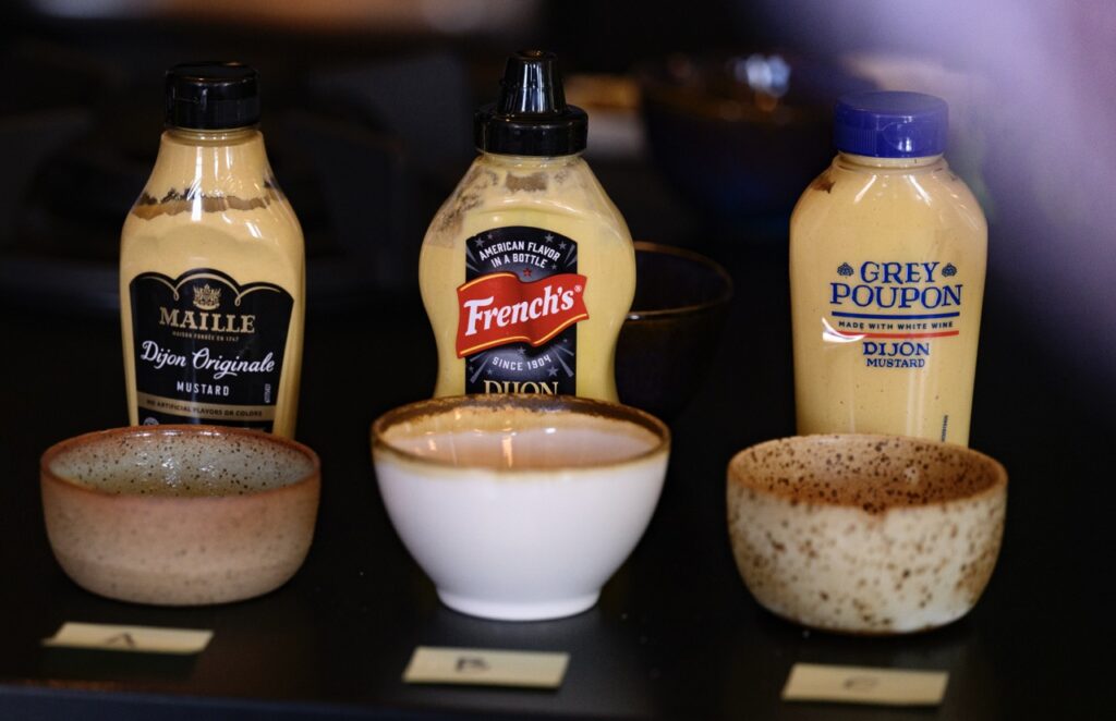 Three bottles of dijon mustard sit in front of three small bowls.
