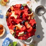 A bow sits on a picnic table filled with star shaped watermelon, strawberries, blueberries, and spinkled with tajin to make a red white and blue fruit salad.