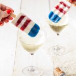 Two people hold red, white, and blue homemade bomb pops above two glasses of clear sparkling liquid on a picnic table.