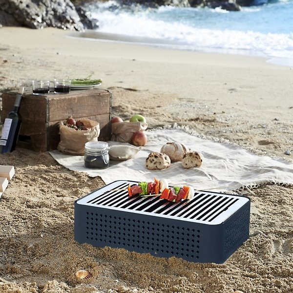 A small blue portable grill sits on the beach with colorful peppers on top with a picnic basket and blanket sitting behind the grill.
