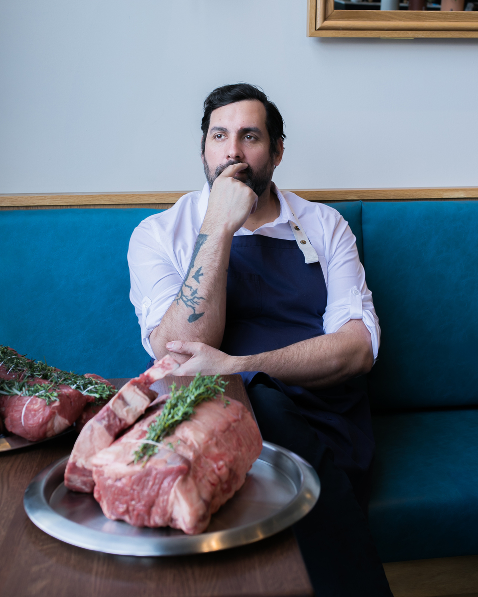 A man sits at a blue booth in a Pittsburgh restaurant with a hand on his chin and a chunk of raw meat on a plate in front of him.