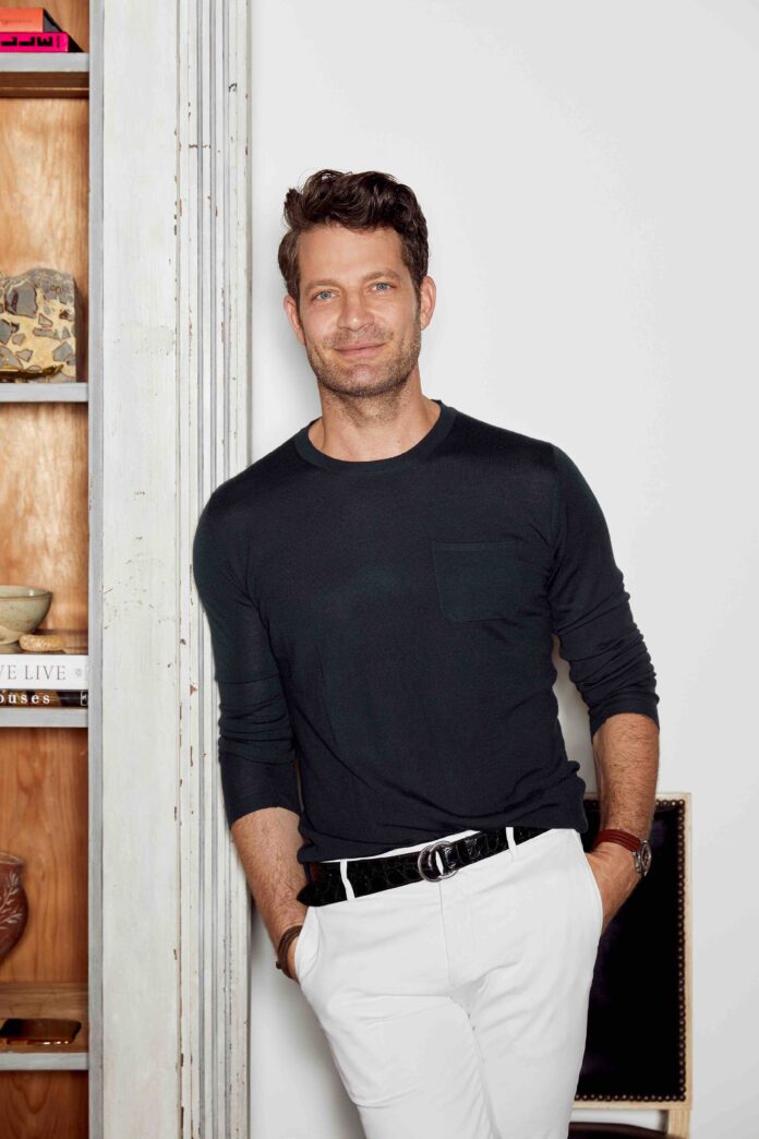 Nate Berkus leans against a white wall in a dark long sleeve shirt and white pants with his hands in his pockets.