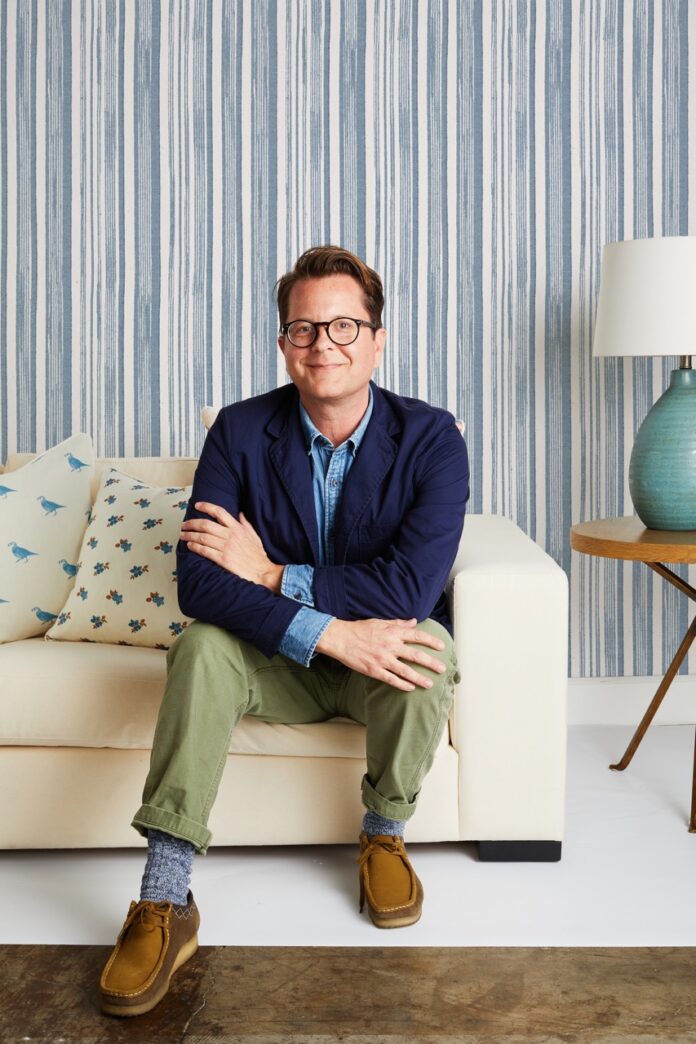 Five Questions With Designer Nathan Turner Nathan Turner sits on a white couch with his arms crossed in a blue cardigan and black glasses.