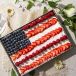 A cake sits in a cake pan decorated with strawberries, raspberries, and blueberries to make a flag design on top.