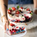 A red, white, and blue gelatin cake decorated with fresh strawberries and blueberries sits on a platter while a woman holds out a cut slice on a small white plate.
