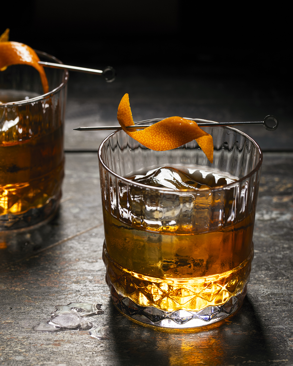 A glass of amber-colored bourbon cocktail garnished with dehydrated orange slices resting on cocktail picks against a dark background with a bottle of bourbon in the background.