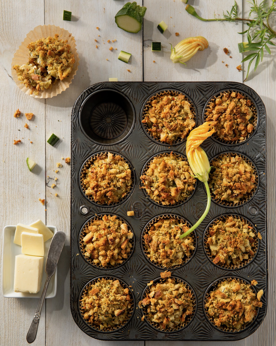 Savory zucchini muffins sit in a muffin tray with a flower over top.