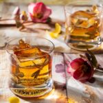 Two small cocktail glasses hold a brown cocktail with dragonflies on the outside of the glasses. Pink flowers are spread across the table.