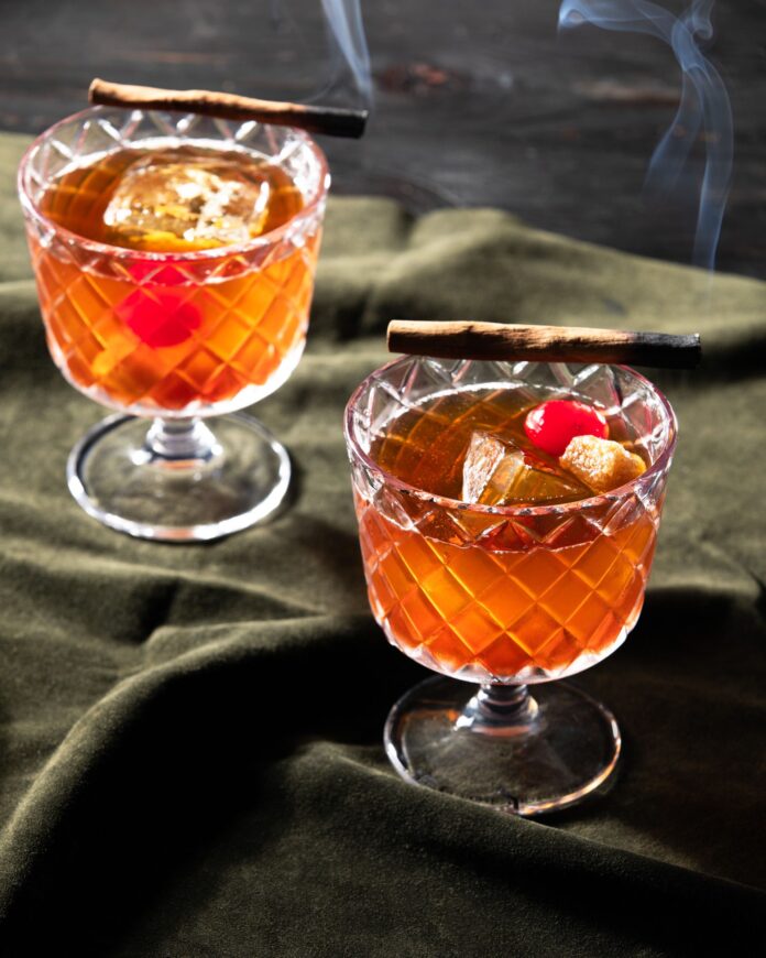 Vandal, A Cocktail for The Bikeriders Two Rocks glasses hold the orange Vandal Cocktail based on The Bikeriders with a sugar cube, cherry, and smoked cinnamon stick garnish. They sit on a green cloth.