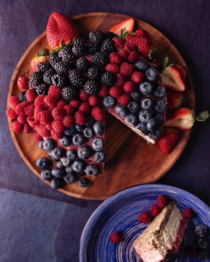 A brown plate holds a vegan cheesecake coated in raspberries, blueberries, and a strawberry swirl.