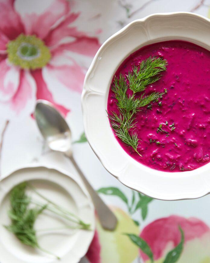 A pink chlodnik soup sits in a bowl with a sprig of green garnish against a pink flower wallpaper.