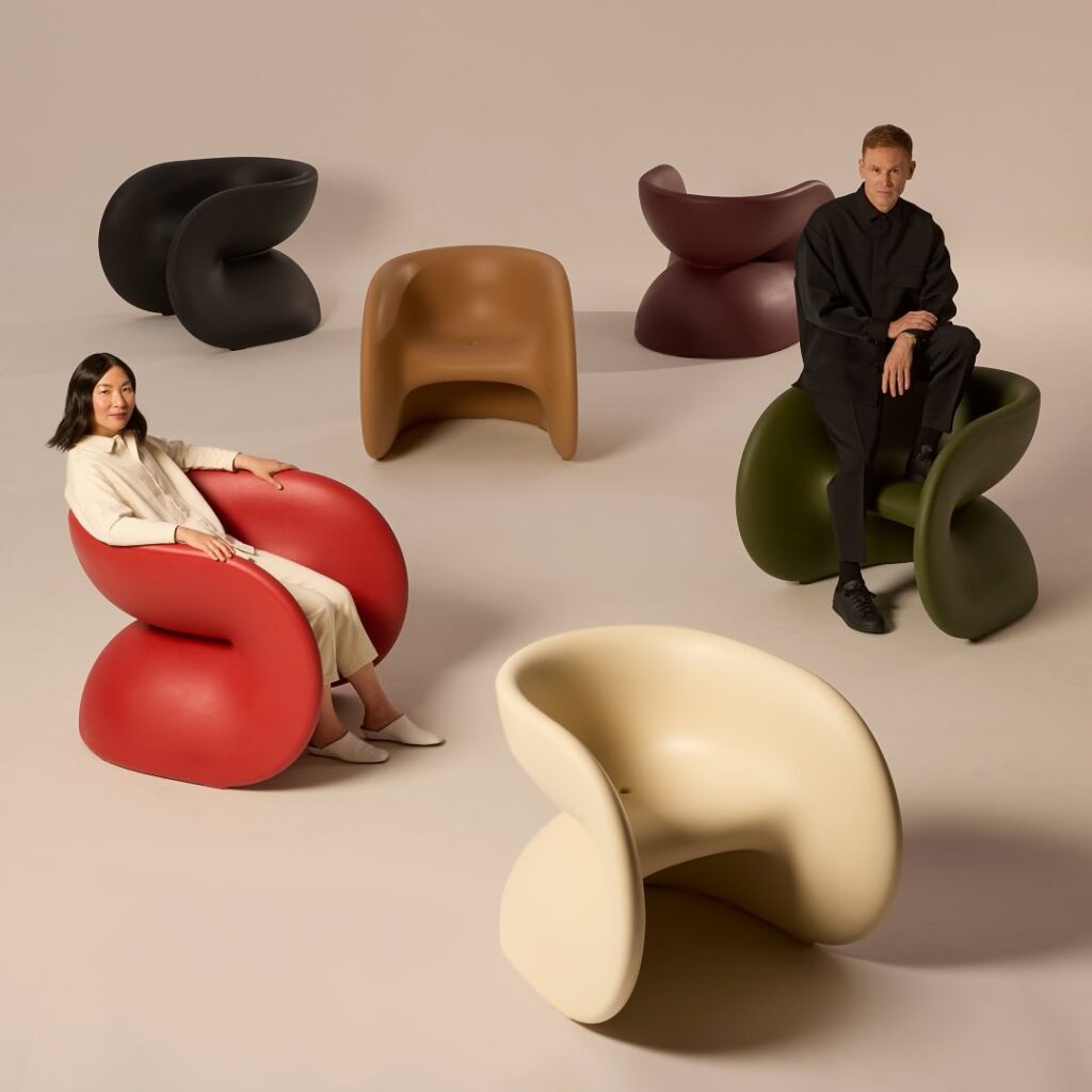 Two people sit in two tubular chairs, one red and one green while other colors of tubular chairs sit amongst the white background.
