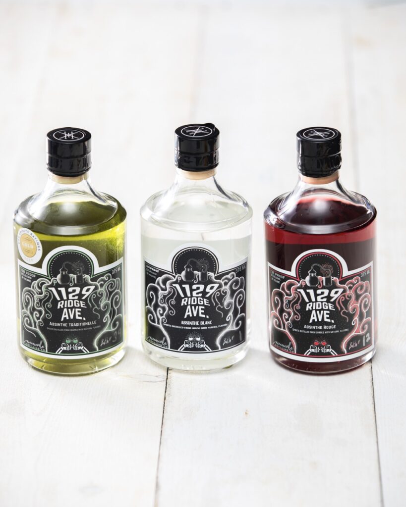 Three bottles of absinthe in green, clear, and red sit on a white picnic table.